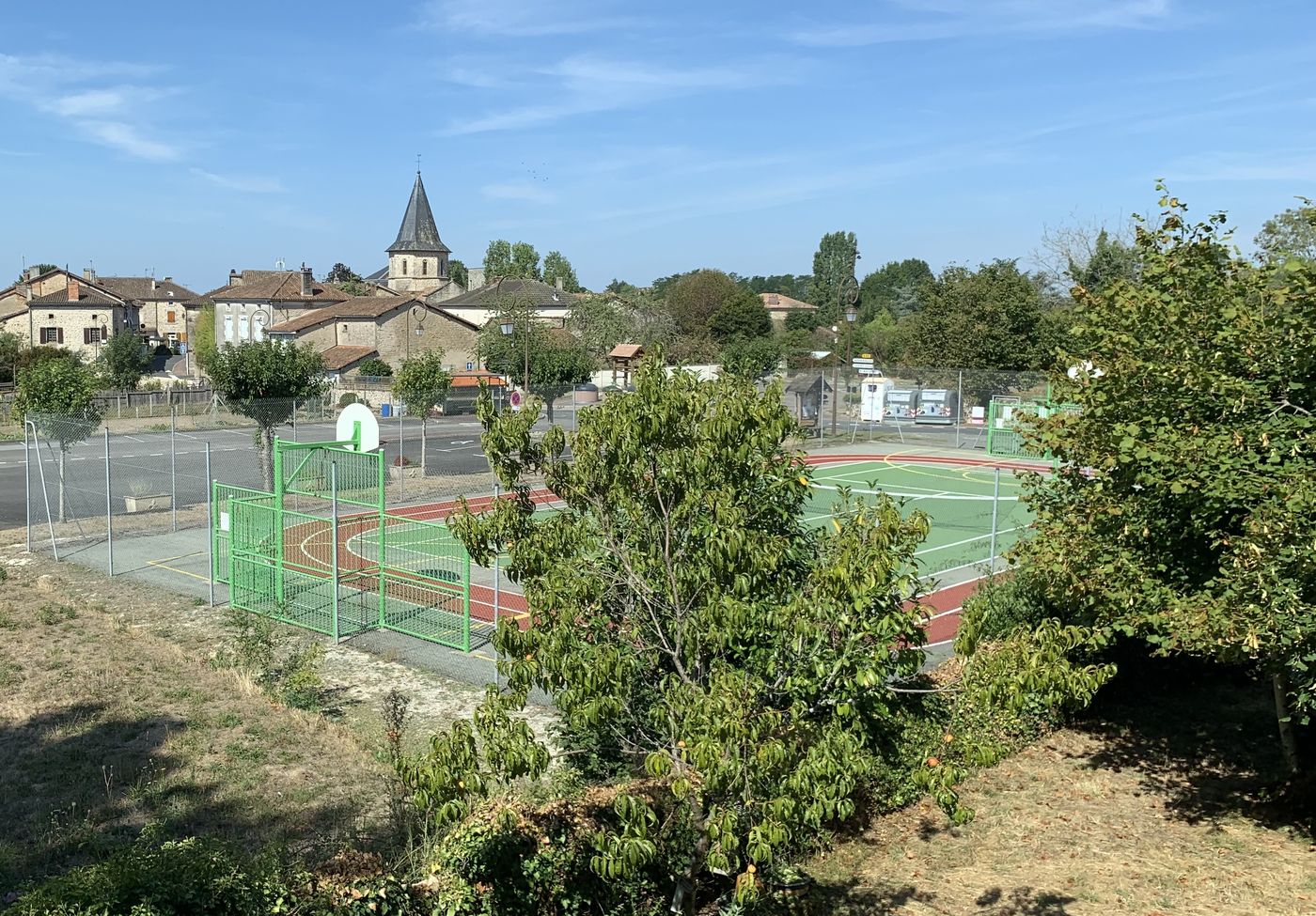 Le terrain multisports du bourg, avec ses paniers de basket ; le clocher de l'église se détache à l'arrière-plan.